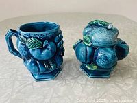 Photo showing both items in set: a coffee mug and lidded sugar bowl, each with raised detailed blue fruit and green leaf accents.