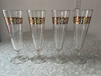 Photo showing front view of four Libbey pilsner glasses lined up with gold laurel leaf pattern on band near rim, standing on a patterned surface.