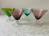 Four differently colored jewel-toned martini glasses with ball stems arranged close together. Colors are green, purple, blue, and smoky purple.