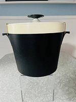 Front view of black and white ice bucket showing white lid with black knob and metal brackets securing lid to base, set on clear stand.
