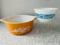 Side-by-side view of two vintage Pyrex bowls: orange Butterfly Gold casserole dish with white floral print and blue Horizon Blue mixing bowl on clear stand