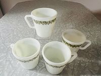 Four vintage Pyrex mugs in white milk glass with green floral band pattern, shown from top angle and side views on table.