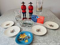 Full lot showing all Canadiana items: two Mountie figurines, four Expo67 pins in original packaging, two Expo67 glasses, glass ashtray, blue floral souvenir dish, and four Centennial dishes with Canadian coat of arms