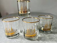 Four vintage Cutler Signature lowball glasses with gold etched ship design and gold rims on grey surface