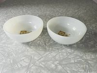 Pair of two white milk glass Fire King bowls with a gold snowflake and starburst design on the interior bottom surface, viewed angled from above on a textured silver surface.
