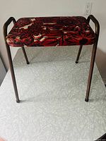 Full view of the vintage stool showing its red and black floral vinyl cushioned seat and metal legs with armrests, on a speckled surface.