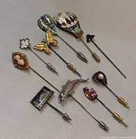Top view of all 10 vintage stick pins arranged side by side on a grey surface, showing various detailed designs and colors.