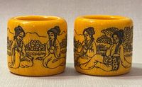 Front view of two yellow ceramic thumb rings decorated with black hand-drawn Asian figures and nature scenes