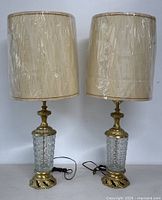 Pair of vintage cut glass and brass table lamps with beige fabric shades. Both lamps are standing upright showing full front view and cords awaiting use.