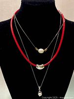 Three necklaces displayed on black bust: red braided leather cord with silver detail, two fine silver chains with different pendants