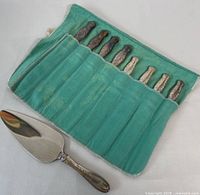 Seven sterling silver handle cutting tools with ornate detailing stored in a green felt fold-up pouch, plus a sterling silver handle cake server placed beside it on a flat surface.