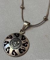 Close-up of the Aztec-themed round pendant showing engraved sun face design and black enamel details on a silver base.