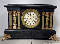 Front view of antique Seth Thomas mantel clock showing black body with four cream-colored pillars, gold lion head columns and feet, and white Roman numeral clock face.