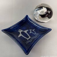 Blue square-shaped glass trinket dish with white penguin designs beside clear round glass penguin paperweight.