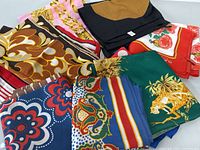 Photo showing several folded scarves with different colors and patterns including pink chain design, brown abstract, blue floral, green tiger print, black, and red scarf.