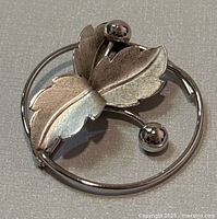 Close-up image of the silver botanical brooch showing the two textured leaves and three spheres on stems with a circular frame.