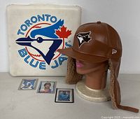 Brown leather-style winter hat with faux fur lining and ear flaps, wearing a mannequin head, with a Toronto Blue Jays branded square cushion and three Roberto Alomar baseball cards co-displayed.