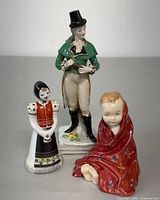Front and side view of three porcelain figurines on white surface: Royal Doulton man figurine, Hungarian woman figurine, and child in red blanket figurine.