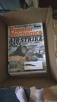 Photo showing 12 Popular Mechanics magazines stacked inside a cardboard box, with the top issue titled Air Strike 2025 featuring military aircraft on the cover.