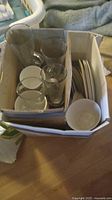 Photo showing a cardboard box filled with various white dishes - plates and bowls, alongside clear glasses of different sizes including mugs and drinking glasses.