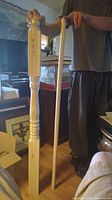 Two wooden pieces held vertically by a person: a carved railing post with a square base and a smooth dowel rod.