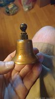 Handheld small brass Liberty Bell replica with black wooden handle shown from front
