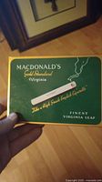 Front side of MacDonald's Gold Standard Virginia cigarette tin showing cigarette image, branding, and slogan.