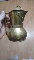 Photo of vintage brass creamer held upright, showing hammered and smooth brass body with handle and lid.