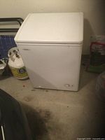 Front and top angled view of a white compact chest freezer named Danny, placed on a floor next to a propane tank and some bags.