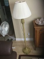 Full view of tall gold metal floor lamp with white pleated fabric shade standing on floor near a wall and furniture.