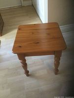 Small wooden square table shown full view with light brown varnish finish and turned legs on a hardwood floor.