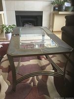 Photo showing modern coffee table with bronze metal frame and clear rectangular glass top, angled legs, geometric cross supports.
