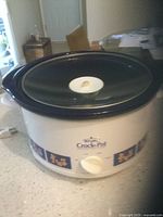 Photo of Rival Crock-Pot slow cooker with blue floral pattern on white base, black lid with white knob on top, placed on a table inside a room.