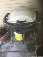 Medium Weber gas grill with white lid and black base on a metal stand. Two propane tanks are placed below the grill. Grill and stand have surface dirt and wear marks.