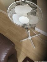 Single white metal pedestal fan on wooden floor showing the protective grille, blades, base, and power cord.