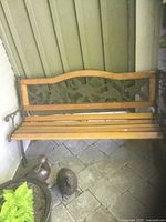 Full view of wooden slatted seat with patterned cast iron and wooden backrest, and cast iron armrests and legs against outdoor siding, shown on stone patio with plants nearby.