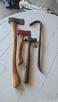 Four hand tools arranged on a white round table: axe, hatchet, ball pein hammer, and crowbar.
