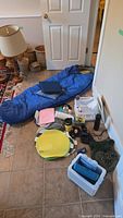 Wide photo showing the blue Taiga sleeping bag on the floor spread out with camping plates, binoculars, cooler, and other small camping accessories around it.