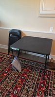 Black folding card table set against wall, folded chairs stacked next to it, set on patterned rug.