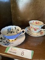 Two tea cup and saucer sets on a wooden surface: Royal Stafford blue and black floral, Royal Albert Gaity pink rose floral