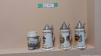 Four German porcelain beer steins arranged in a corner with light peach wall background. Three have pewter lids and detailed crests; fourth has black and white cityscape illustration and no lid.
