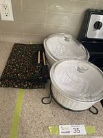 Two white ceramic casserole dishes with glass lids placed on a kitchen counter next to a floral-patterned carrying bag and chopsticks. Dishes have no chips or cracks.