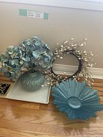 Blue vase holding faux blue and cream hydrangea flowers, blue bowl with scalloped edges, and brown twig wreath with white berries against wall and floor