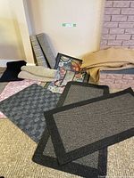 Photo showing a variety of floor mats and runners including two small black border mats, rolled rugs in beige and tan, pink bath mat, floral framed mat, and underlay rolls.