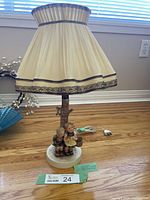 Full view of the vintage Hummel Goebel lamp with cream pleated lampshade edged in brown trim and figurines base.