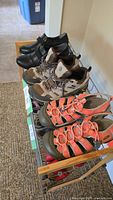 Three pairs of lady's shoes arranged in a row on a shoe rack: black leather casual shoes, beige Merrell hiking sneakers, orange Keen sports sandals.