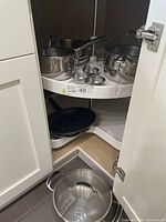 View inside corner kitchen cabinet holding Lagostina stainless steel pots and pans, JA Henckels frying pan, and poached egg cups.