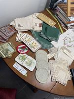 Photo showing a collection of embroidered linen napkins, green quilted napkins, crocheted and ceramic coasters, and a small floral embroidered tablecloth, spread on a wooden table.