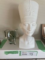 Front view of white ceramic bust of Nefertiti next to clear glass elephant paperweight on shelf.