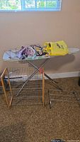 Ironing board with metal legs and fabric cover, assorted clothes hangers on top, and shoe shelves on floor beneath.